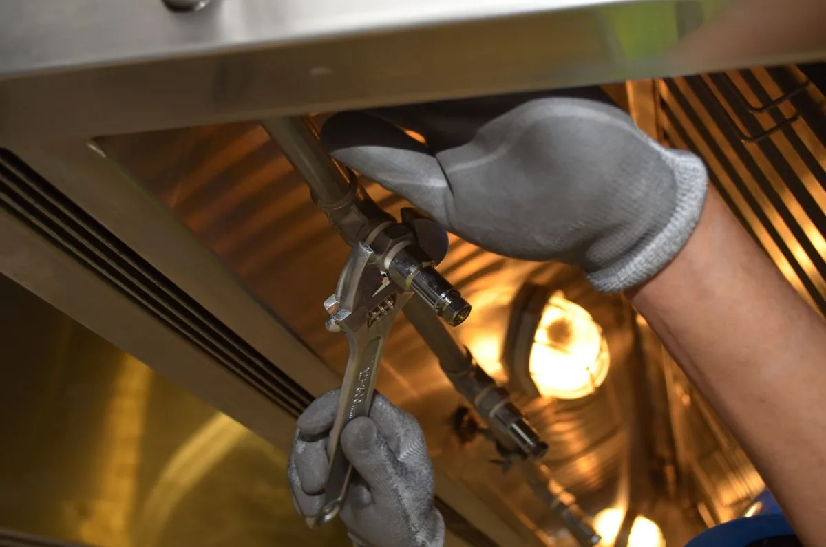 Mãos de um técnico com luvas apertando uma conexão com chave inglesa num equipamento de cozinha industrial